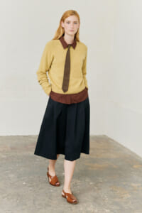 Woman modeling a layered preppy outfit: tan knit sweater over brown shirt with a tie, black midi skirt, and brown sandals.