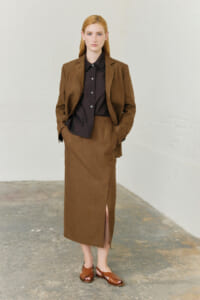 Woman in a brown blazer with matching skirt and black button-down shirt, hands in pockets against a light wall.