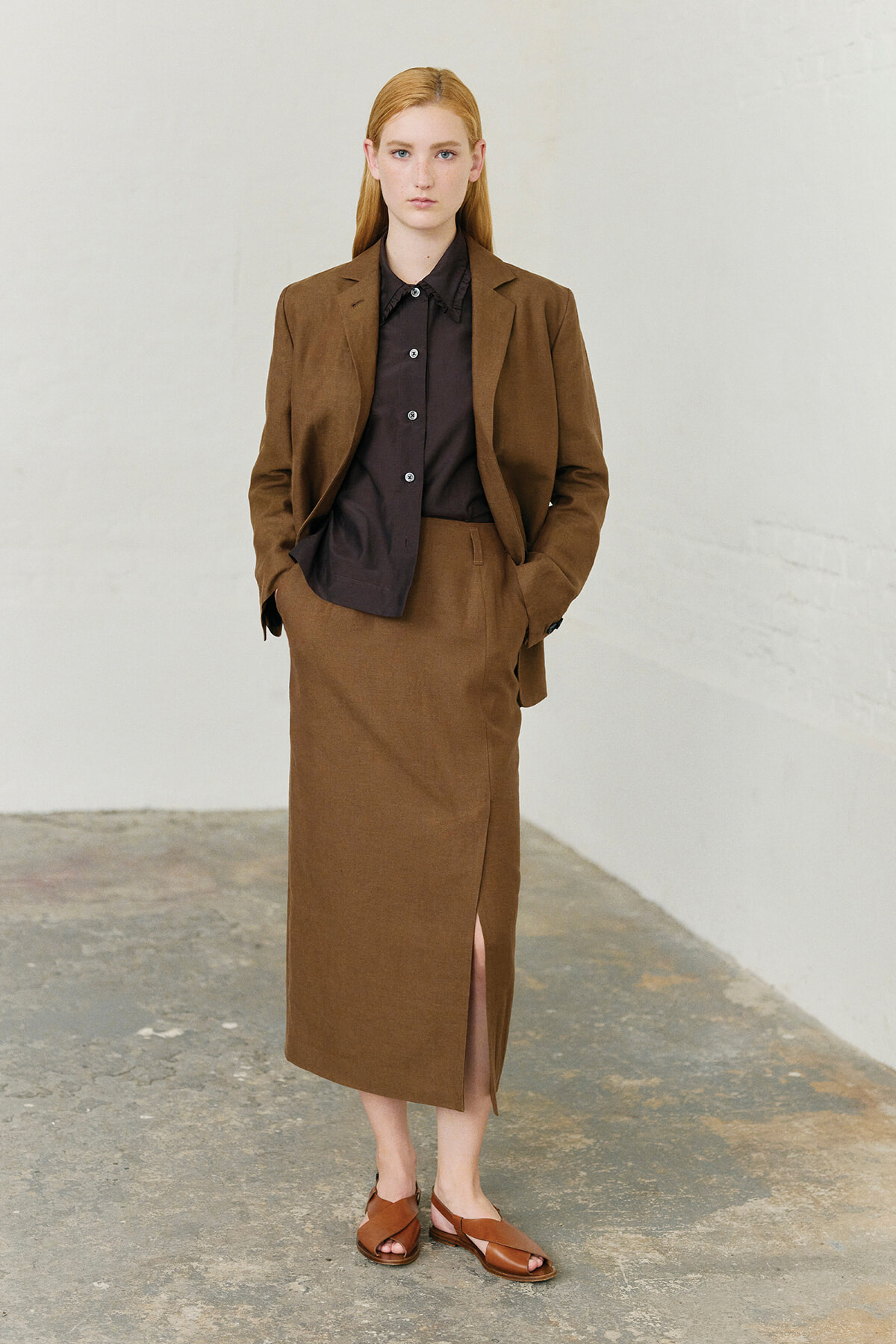 Woman in a brown blazer with matching skirt and black button-down shirt, hands in pockets against a light wall.