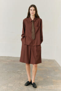 Model in a brown matching shirt and knee-length shorts, brown tie, black loafers, standing against a light wall.”