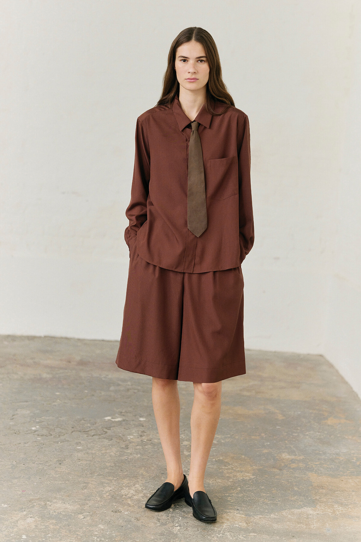 Model in a brown matching shirt and knee-length shorts, brown tie, black loafers, standing against a light wall.”
