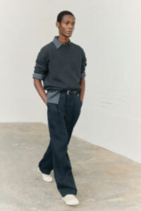 Male model walking in a dark gray sweater layered over a gray collared shirt, navy trousers, and white sneakers.