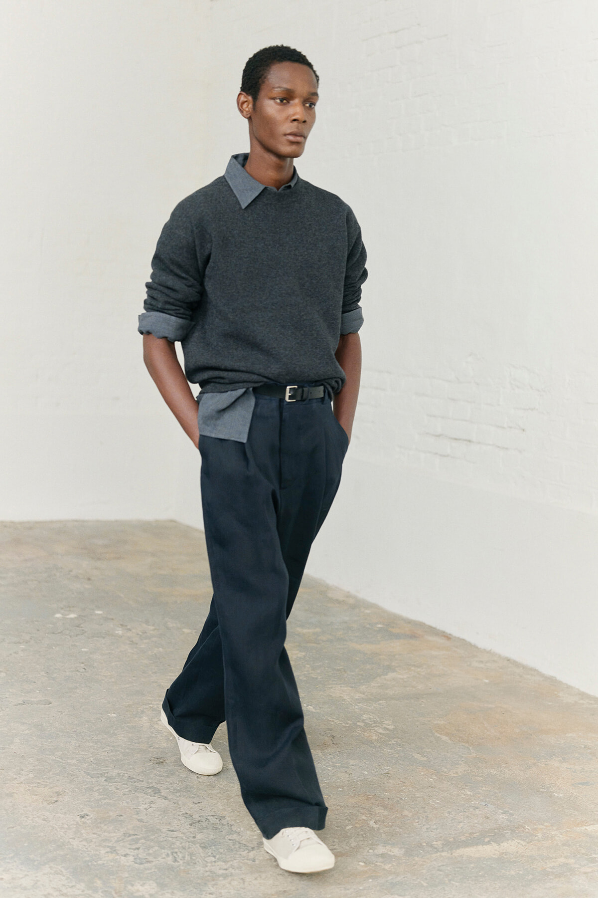 Male model walking in a dark gray sweater layered over a gray collared shirt, navy trousers, and white sneakers.