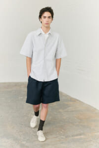 Young man wearing a light oversized short-sleeve shirt, navy shorts, black socks, and white sneakers in a minimalist studio setting.