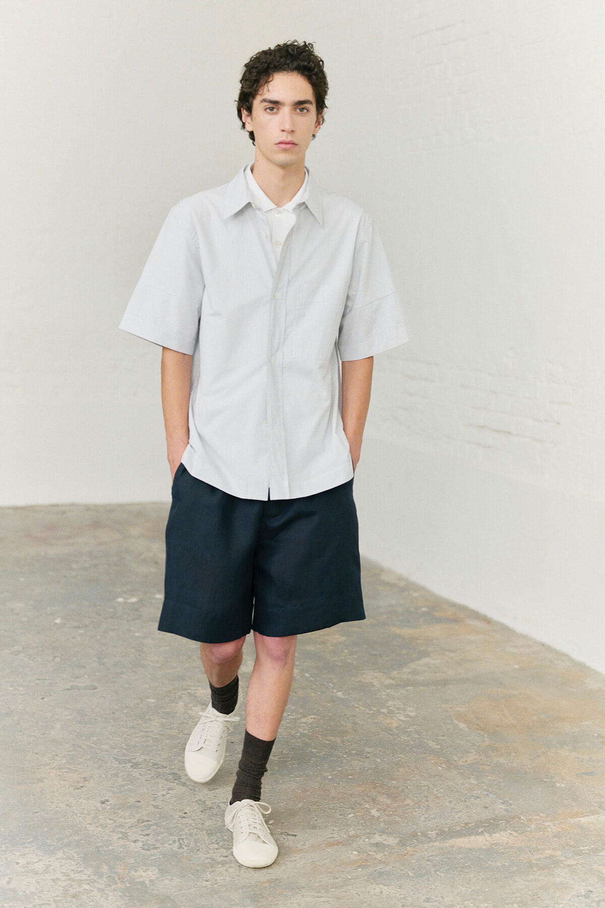 Young man wearing a light oversized short-sleeve shirt, navy shorts, black socks, and white sneakers in a minimalist studio setting.