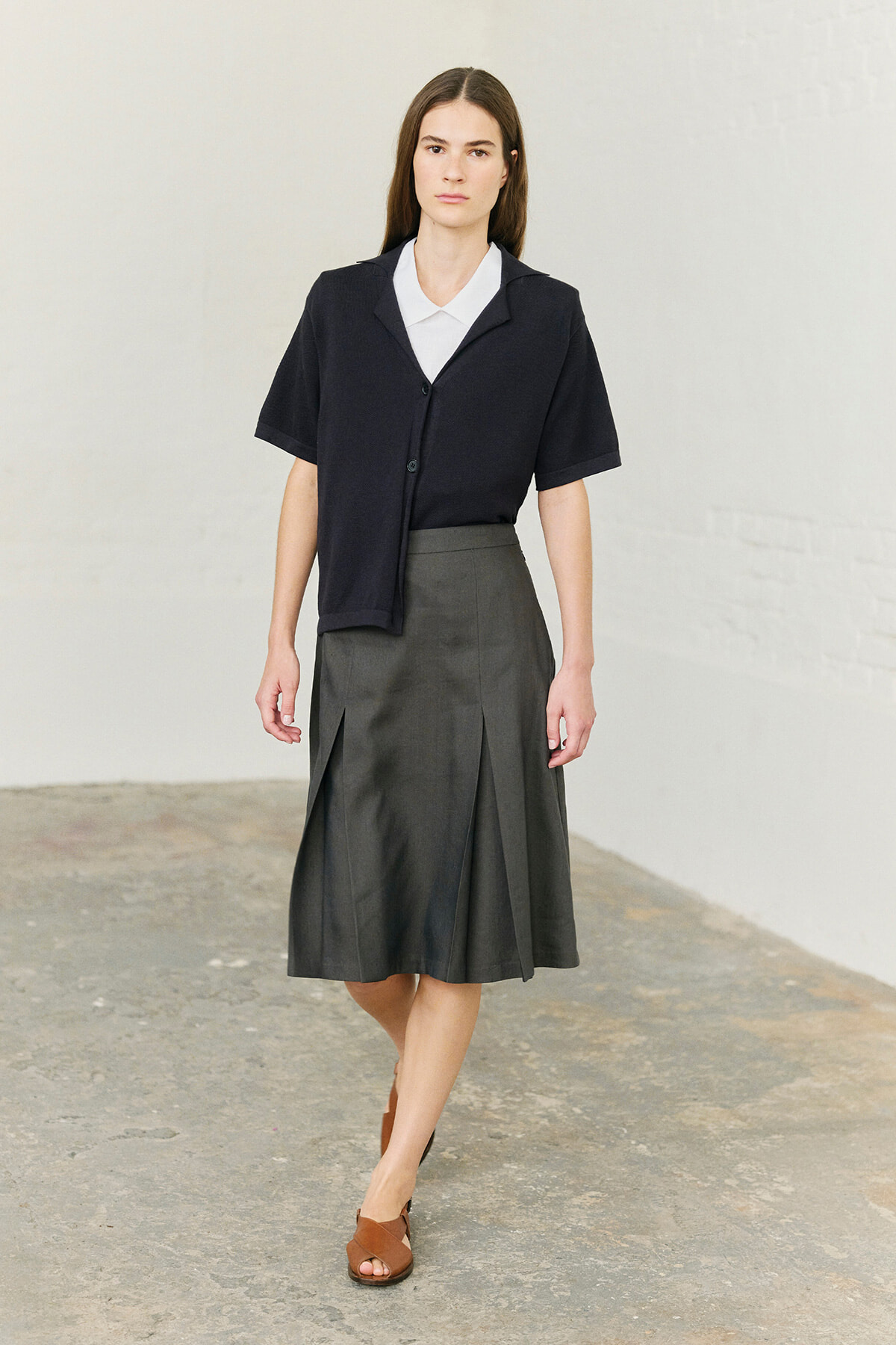 Woman wearing a navy short-sleeve cardigan over a white collared shirt and a gray pleated midi skirt, standing in a minimalist studio.