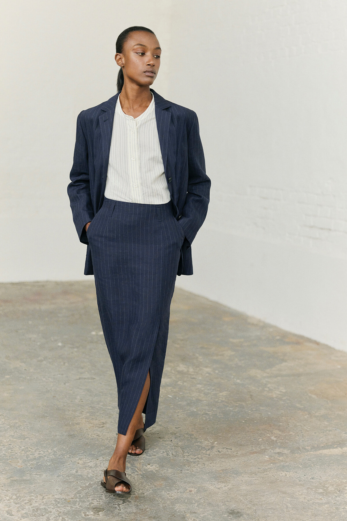 Model wearing a navy pinstripe suit (blazer and pencil skirt) with a white blouse, walking toward the left in a minimalist space.