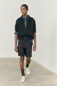 Young man in a blue shirt with a tan tie, dark zip sweater, navy shorts, and white sneakers walking in a light, blank setting.