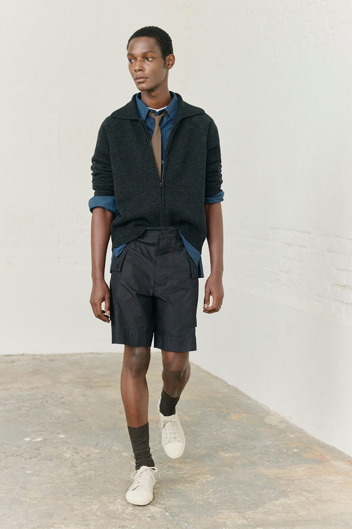 Young man in a blue shirt with a tan tie, dark zip sweater, navy shorts, and white sneakers walking in a light, blank setting.