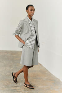 Woman modeling a light gray blazer and knee-length shorts with a plaid button-down shirt, standing with hands in pockets against a neutral background, minimalist studio.