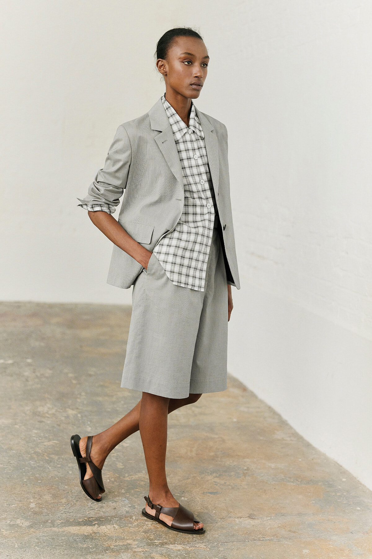 Woman modeling a light gray blazer and knee-length shorts with a plaid button-down shirt, standing with hands in pockets against a neutral background, minimalist studio.