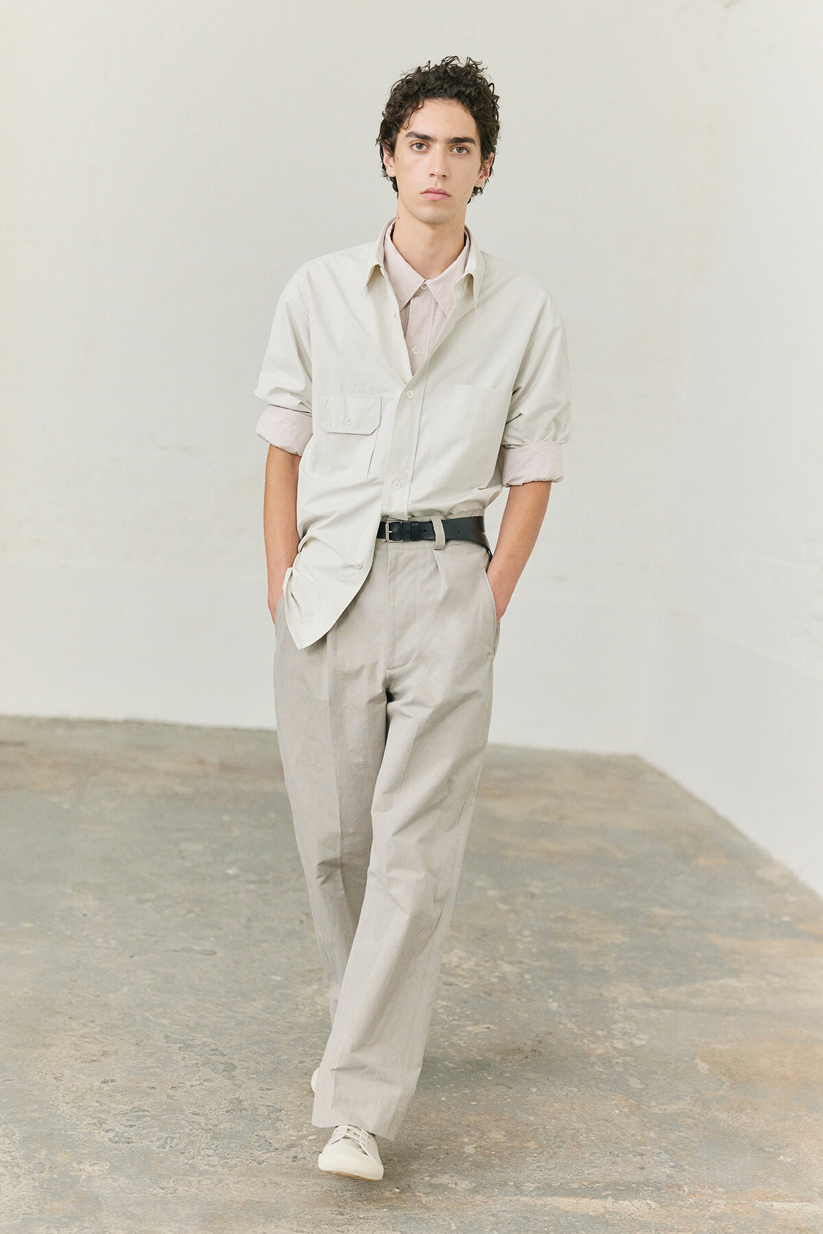 Young man in a beige, casual suit walking toward the camera in a minimalist studio setting with hands in pockets.