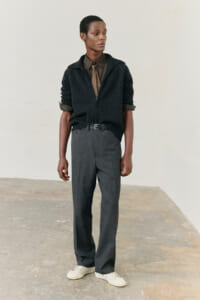 Full-body portrait of a man in smart-casual attire: dark zip-up sweater over an olive shirt with a brown tie, gray trousers, and white sneakers, standing against a pale wall.