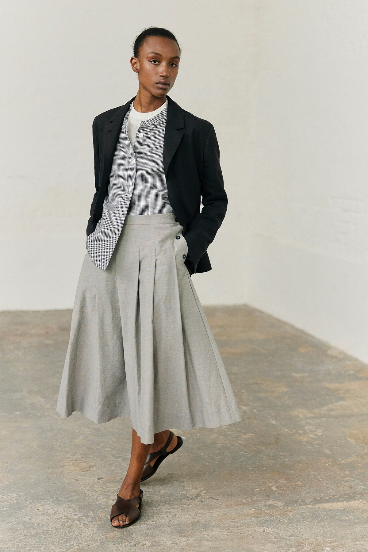 Person standing in a neutral space wearing a black blazer over a light checkered top and a full, light gray midi skirt with sandals.