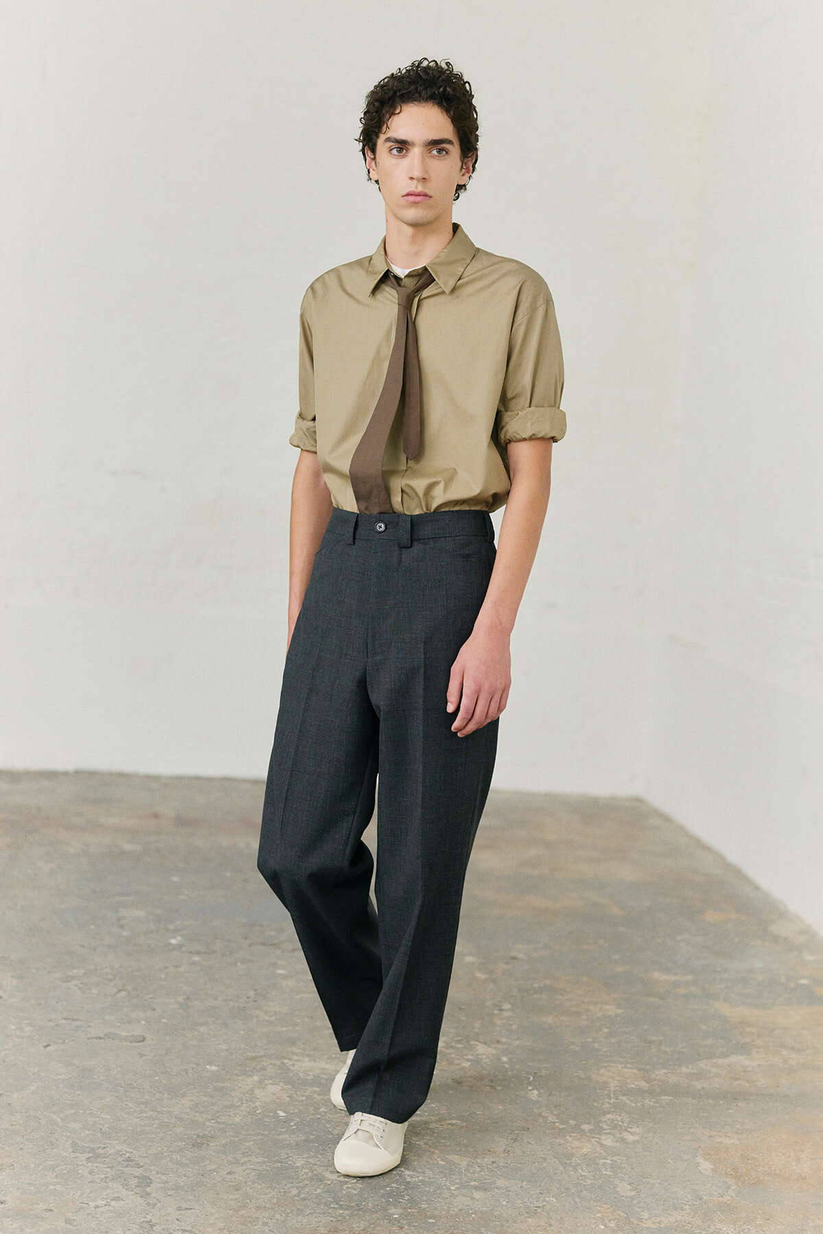 Male model in a tan button-up shirt with rolled sleeves, a loosely tied brown tie, and dark gray wide-leg trousers in a neutral studio.
