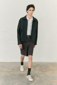 Young man in a dark green jacket over a light shirt, with dark shorts and white sneakers, posing in a neutral studio setting.