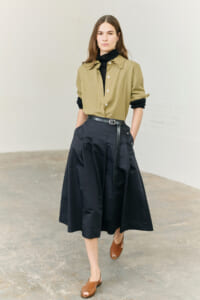 Model wearing an olive button-down shirt tucked into a black midi skirt with a belt, standing in a neutral studio setting.