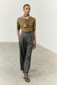 Model wearing a camel knit top with a polka-dotted scarf, gray high-waisted pleated trousers, and brown sandals in a studio setting in a full-length pose.