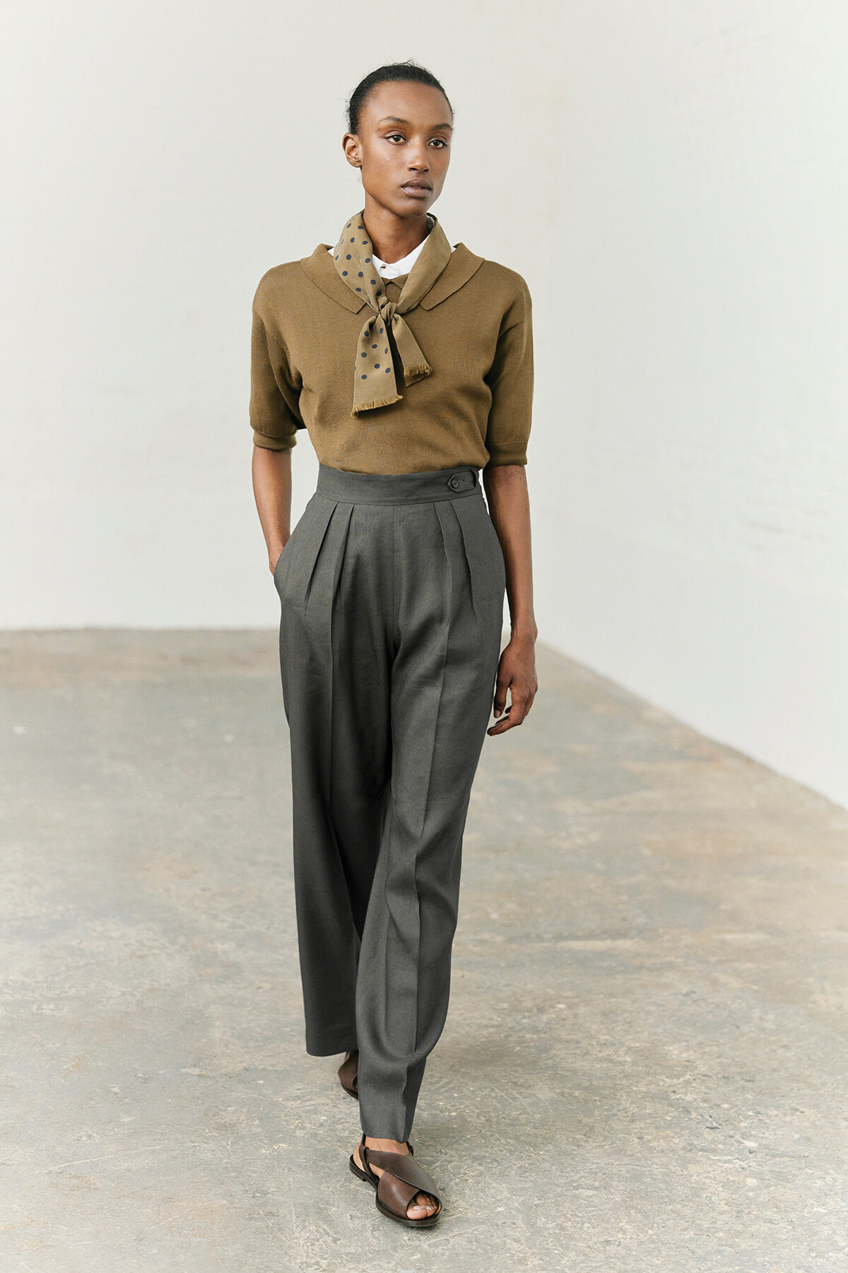 Model wearing a camel knit top with a polka-dotted scarf, gray high-waisted pleated trousers, and brown sandals in a studio setting in a full-length pose.