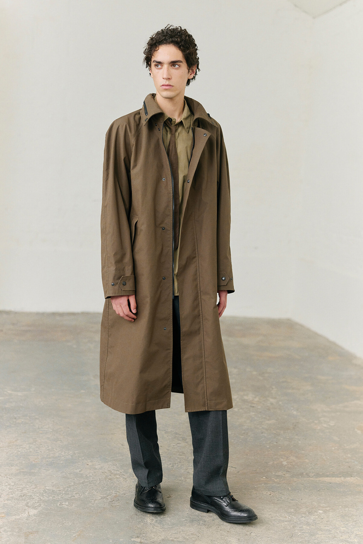 Tall man in a long brown trench coat standing against a light wall, hands at sides, looking off-camera to the left.