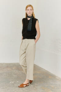 Full-length shot of a woman wearing a black sleeveless cardigan, beige high-waisted trousers, brown sandals, and a green polka-dot scarf.