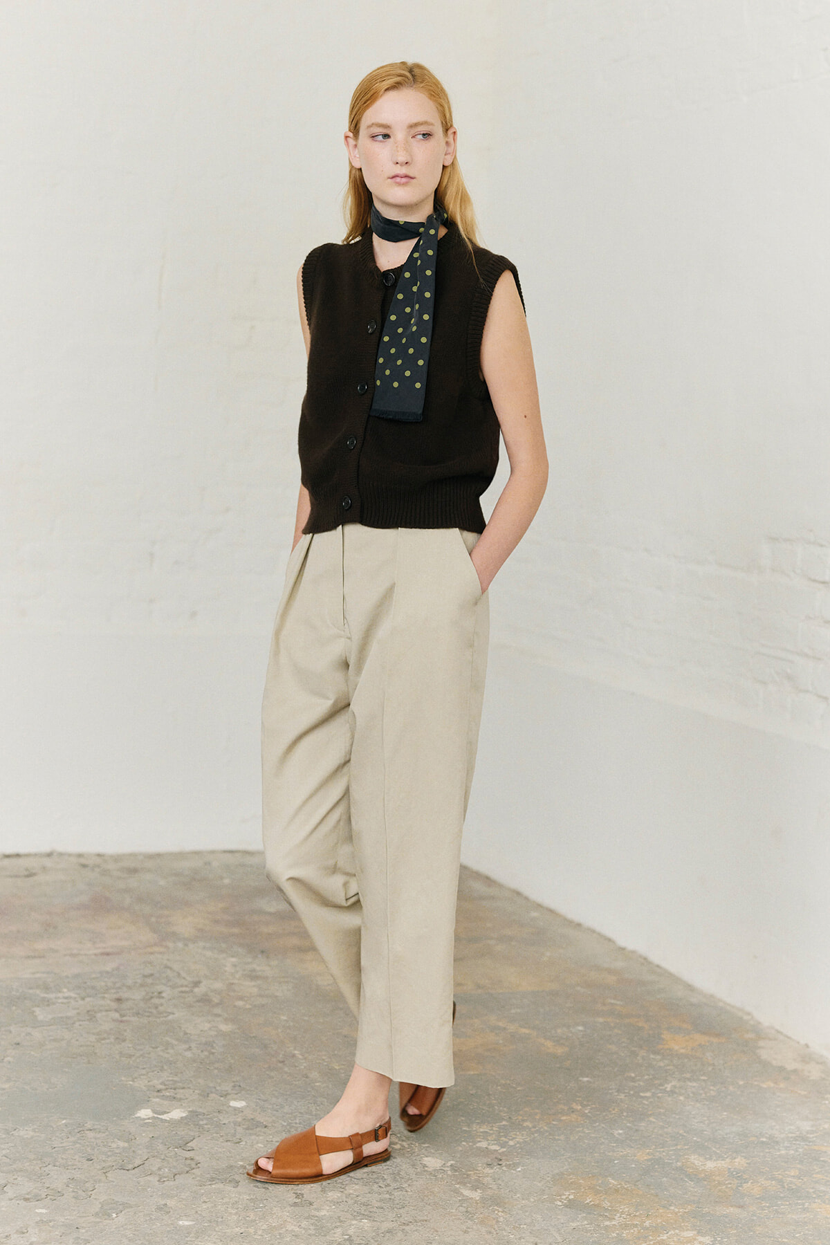 Full-length shot of a woman wearing a black sleeveless cardigan, beige high-waisted trousers, brown sandals, and a green polka-dot scarf.