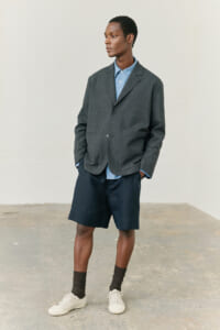 Man wearing a charcoal blazer over a light blue button-down shirt, navy shorts, dark socks, and white sneakers, standing with hands in pockets against a plain backdrop.