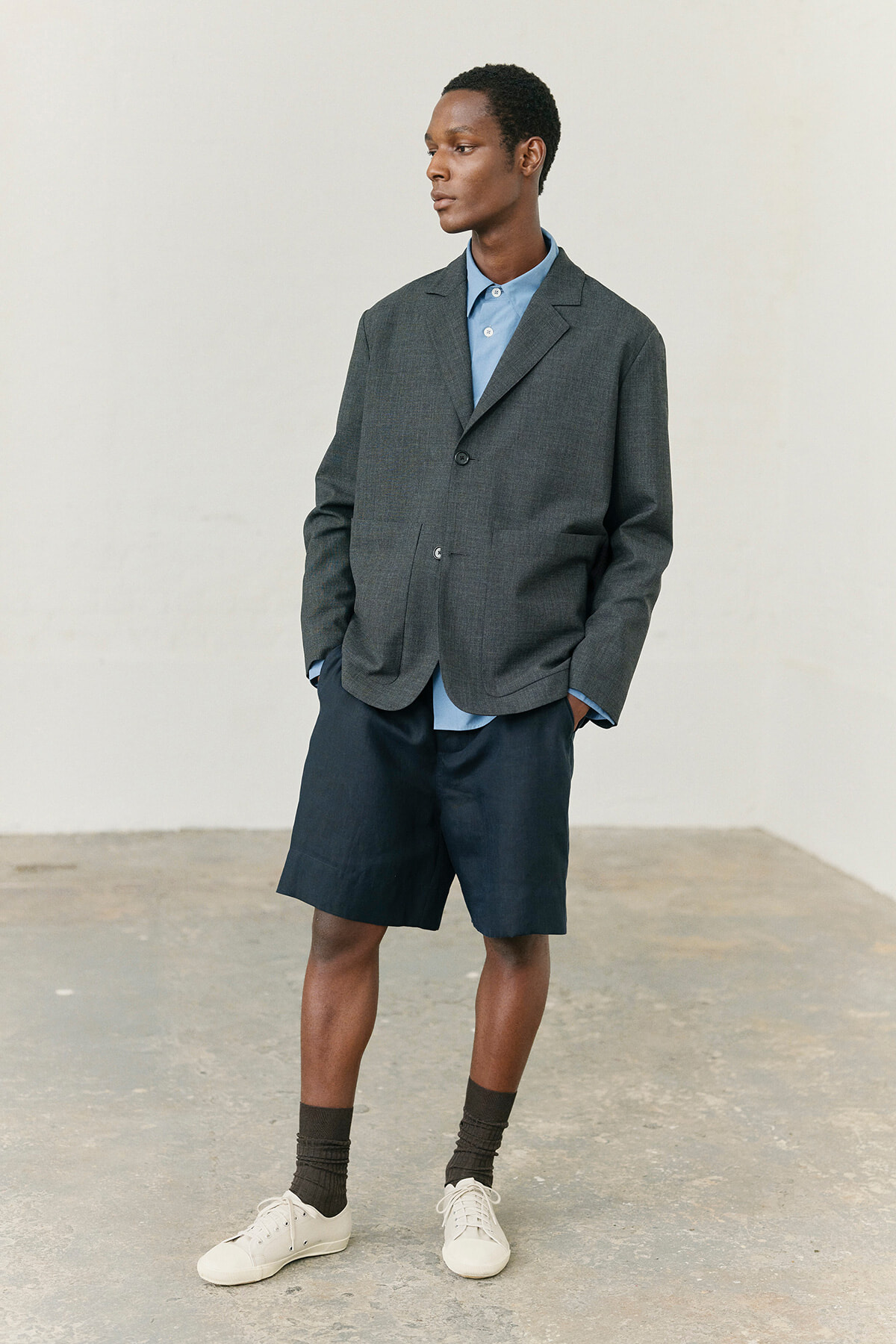 Man wearing a charcoal blazer over a light blue button-down shirt, navy shorts, dark socks, and white sneakers, standing with hands in pockets against a plain backdrop.