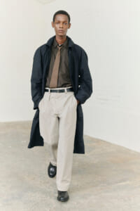 Man wearing a dark trench coat over a brown shirt and tan tie, beige trousers, and black shoes, walking toward the camera with hands in pockets.