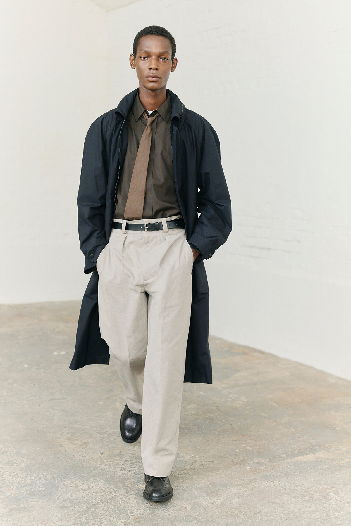 Man wearing a dark trench coat over a brown shirt and tan tie, beige trousers, and black shoes, walking toward the camera with hands in pockets.