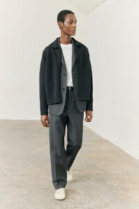 Man wearing a charcoal cardigan over a light gray suit with a white T-shirt, walking in a simple indoor space.