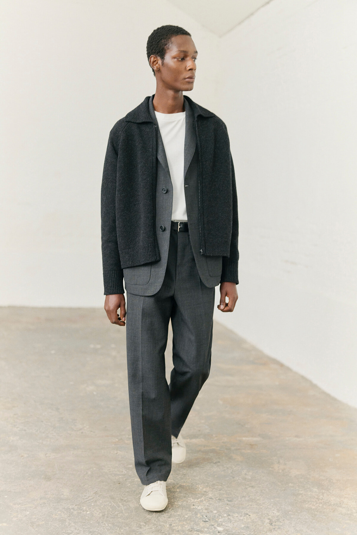 Man wearing a charcoal cardigan over a light gray suit with a white T-shirt, walking in a simple indoor space.