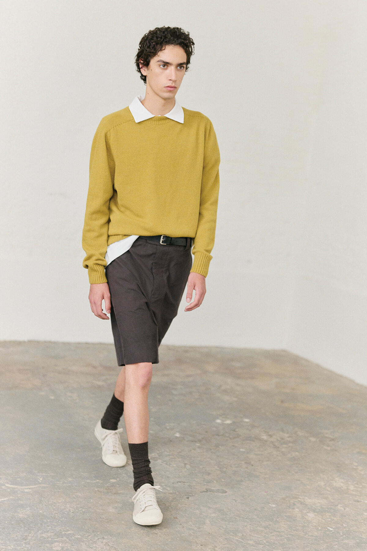 Male model wearing a mustard knit sweater over a white collared shirt, dark knee-length shorts, black socks, and white sneakers, walking in a studio setting.