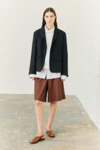 Model wearing a dark blazer over a light knit sweater and white shirt, paired with brown knee-length shorts and brown sandals, standing in a neutral studio.