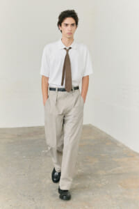 Young man in a white short-sleeve shirt, brown tie, and light gray pleated trousers, standing in a neutral studio background with a black belt and shoes.