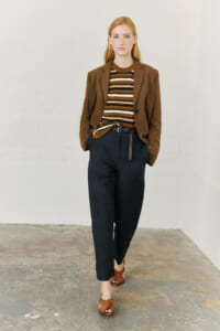 Model wearing a brown blazer over a striped brown, white, and black top with dark tapered pants and brown sandals, walking toward the camera in a white studio.