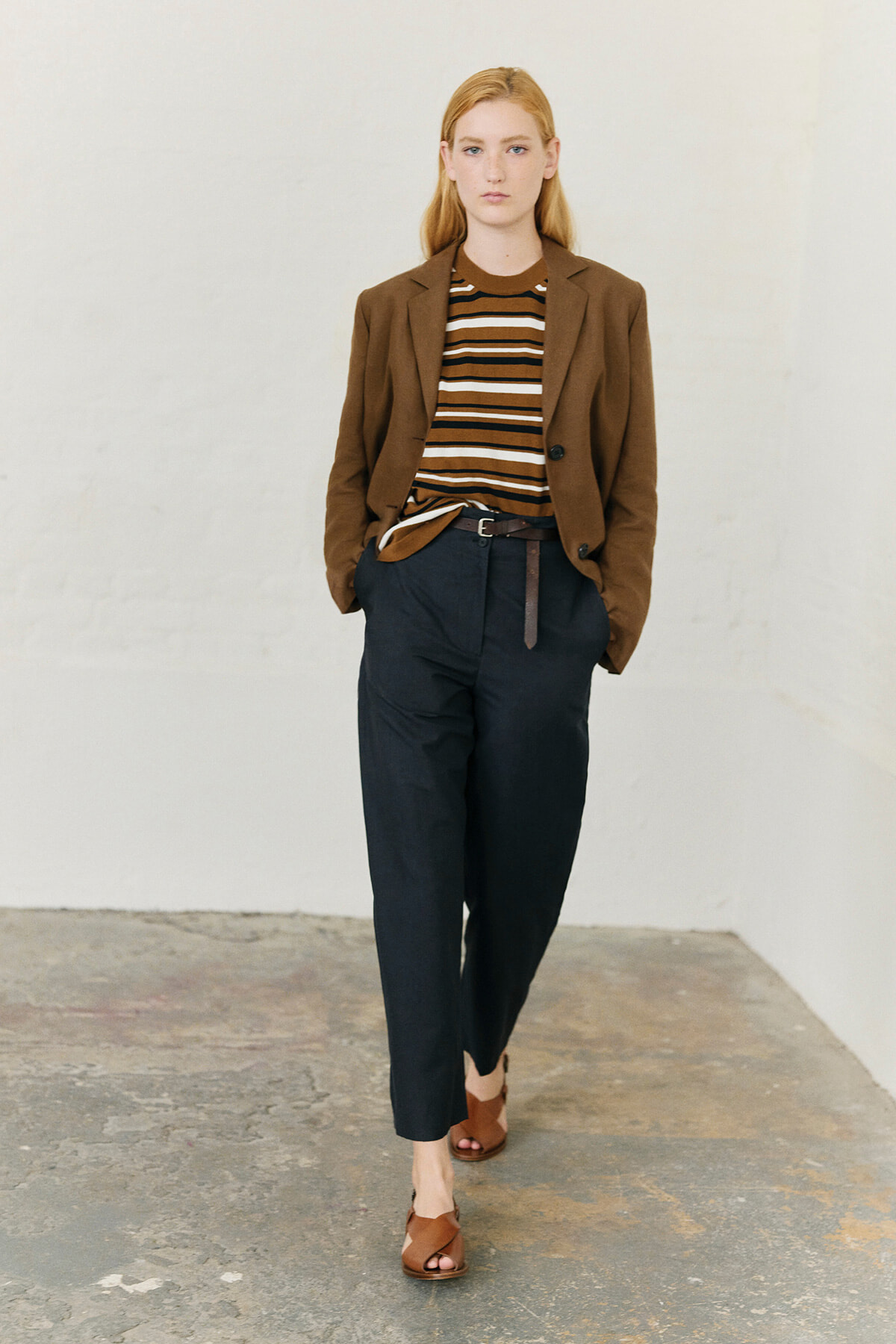 Model wearing a brown blazer over a striped brown, white, and black top with dark tapered pants and brown sandals, walking toward the camera in a white studio.