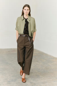 Woman standing in a neutral space wearing a light olive green pinstriped shirt over a dark brown top, high-waisted brown wide-leg pants with a brown belt, and brown sandals.