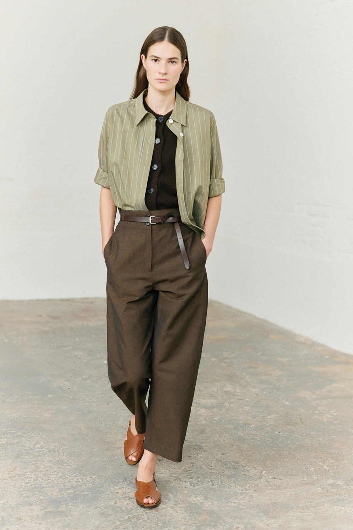 Woman standing in a neutral space wearing a light olive green pinstriped shirt over a dark brown top, high-waisted brown wide-leg pants with a brown belt, and brown sandals.