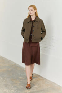 Woman wearing a brown knee-length dress with a matching brown belt detail and an olive bomber jacket, standing against a white brick wall with hands in pockets.