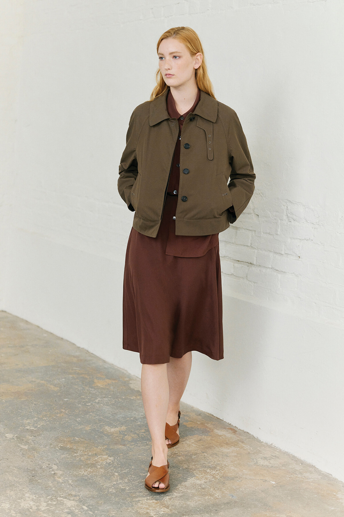 Woman wearing a brown knee-length dress with a matching brown belt detail and an olive bomber jacket, standing against a white brick wall with hands in pockets.