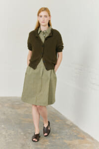 Young woman with blonde hair wearing an olive striped shirt dress and a dark green cardigan, standing with hands in pockets beside a white wall.