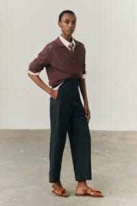 Model wearing a burgundy knit polo over a white shirt with a tie, tucked into high-waisted dark wide-leg pants and brown sandals.