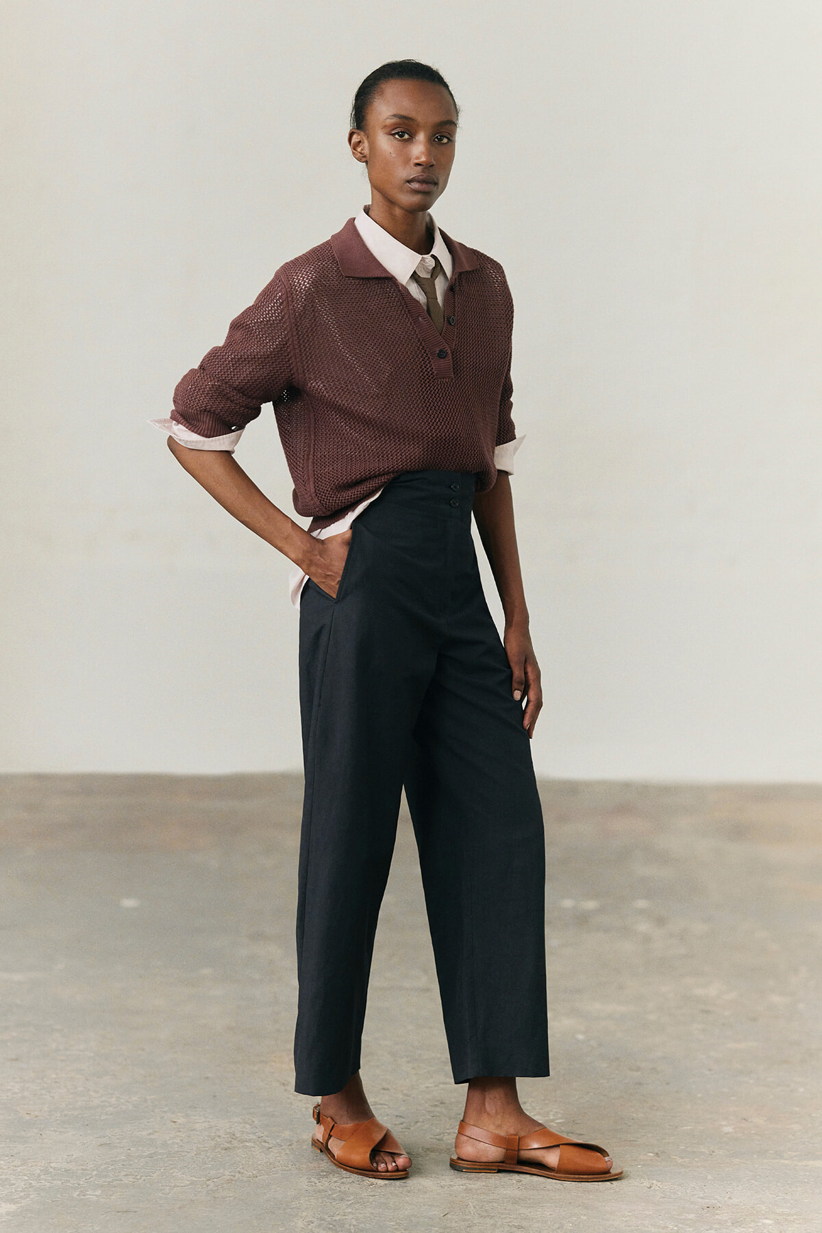 Model wearing a burgundy knit polo over a white shirt with a tie, tucked into high-waisted dark wide-leg pants and brown sandals.