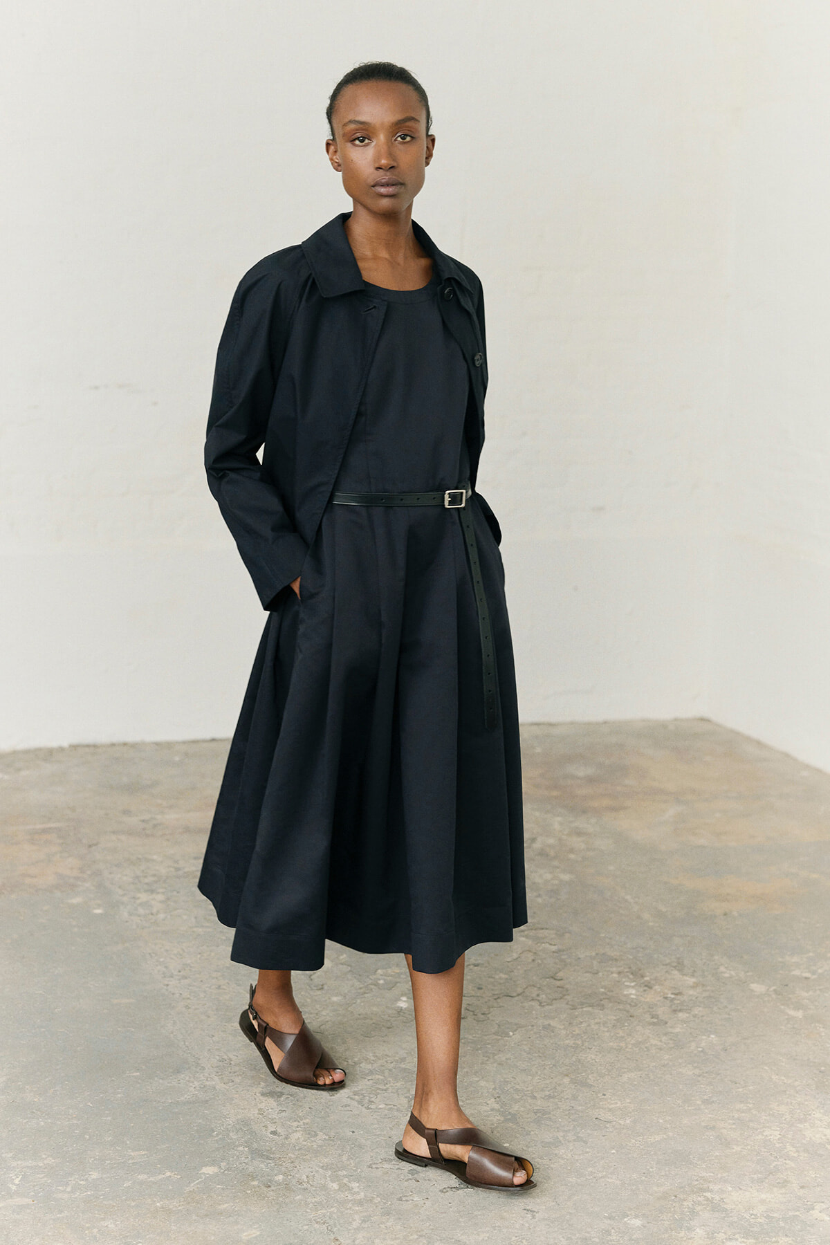 Portrait of a woman in a navy trench-dress with a black belt, hands in pockets, wearing brown sandals, against a light backdrop.
