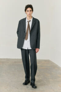 Young man in a dark gray suit with a striped shirt and brown tie, standing in a minimalist studio.