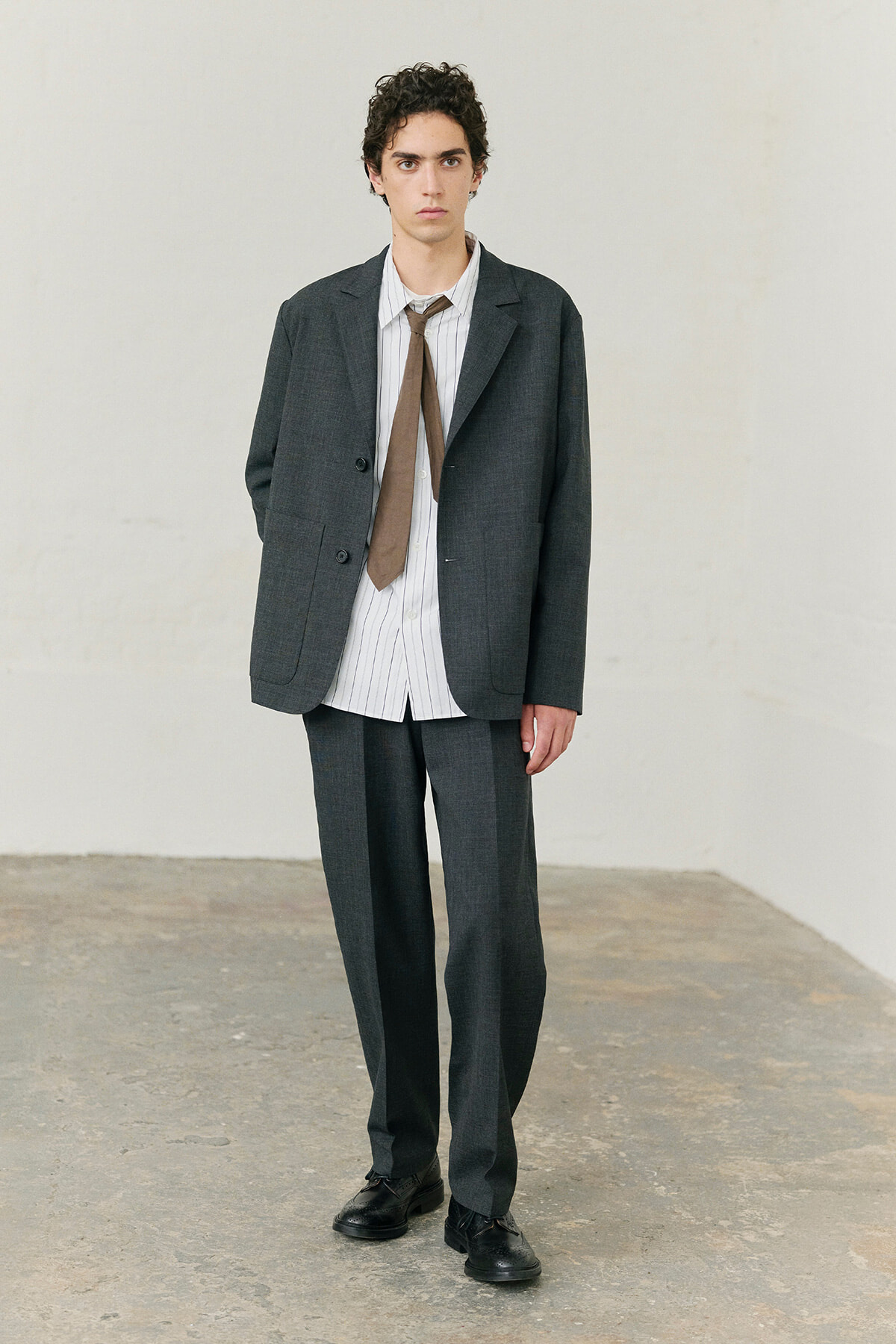 Young man in a dark gray suit with a striped shirt and brown tie, standing in a minimalist studio.