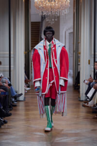 Model walks a runway in an oversized red and white striped coat with blue accents, bright green laced knee-high boots, and a green tie in a grand hallway.