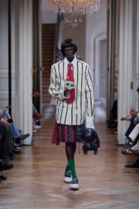 Model on a runway wearing a white striped blazer, red tie, color-blocked pleated skirt, bright green socks, blue sneakers, and carrying a dark plush toy and microphone.
