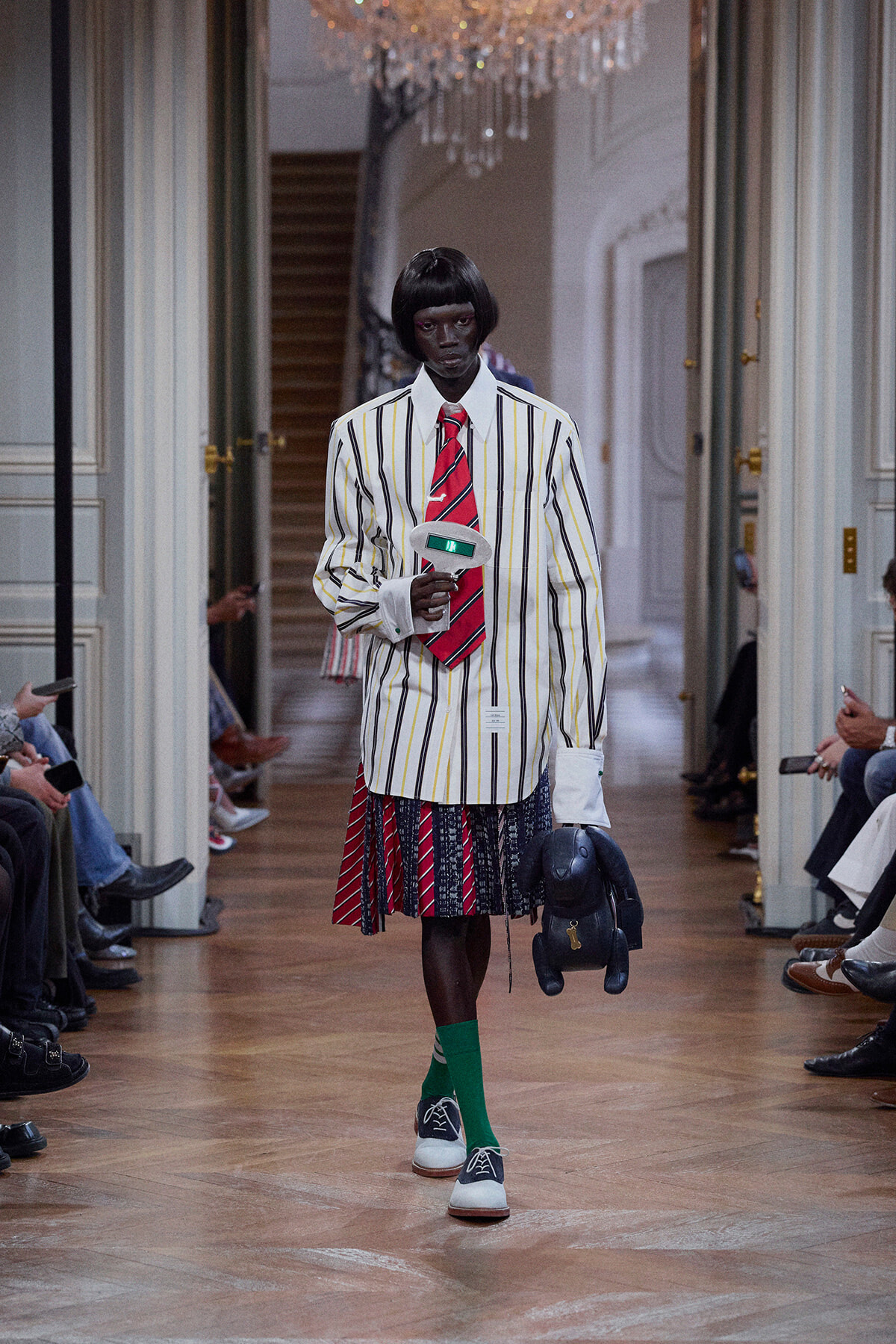 Model on a runway wearing a white striped blazer, red tie, color-blocked pleated skirt, bright green socks, blue sneakers, and carrying a dark plush toy and microphone.
